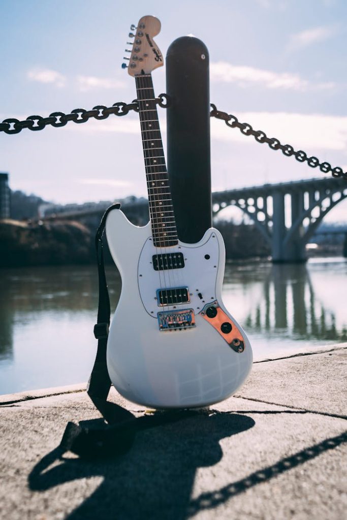 Electric guitar resting by the riverside with a bridge in the background, under daylight.